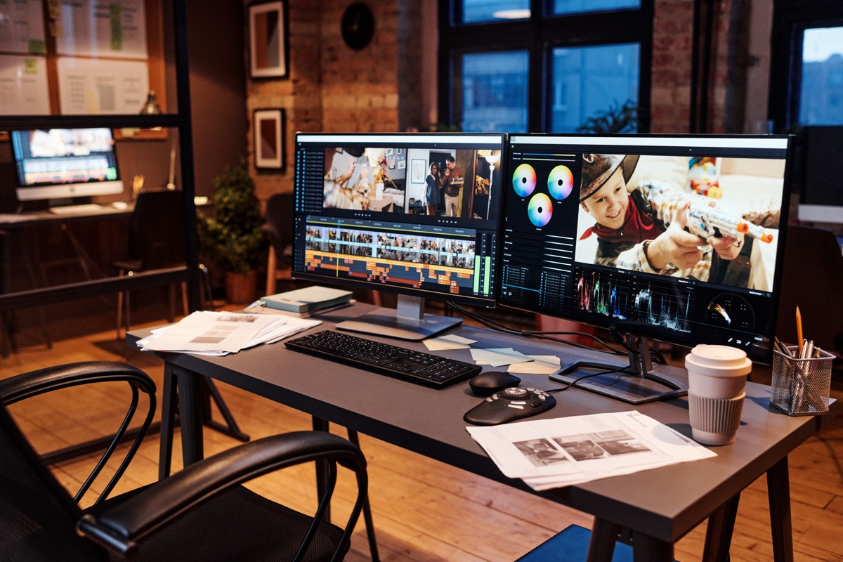 Dual monitors running video editing and colour grading software on a desk in a warm brick-walled office