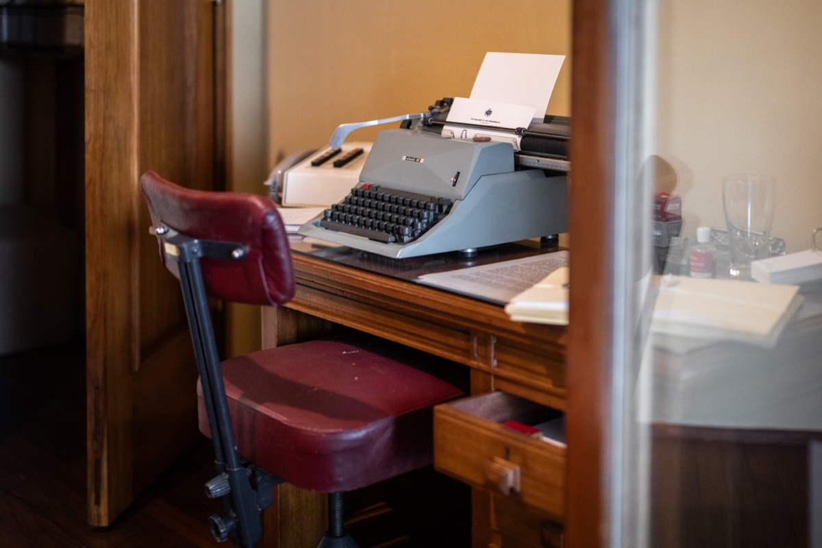 Blue-grey typewriter with paper on wooden desk, burgundy vintage chair tucked underneath