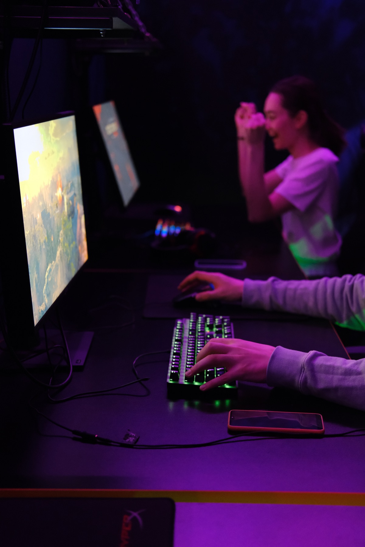 Dark gaming workspace with illuminated monitor showing game scene, RGB keyboard with green backlighting, person gaming