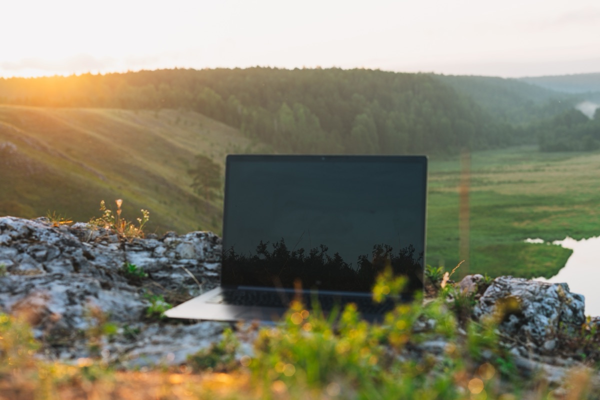 Laptop open on rocky outcrop overlooking green valley at sunset