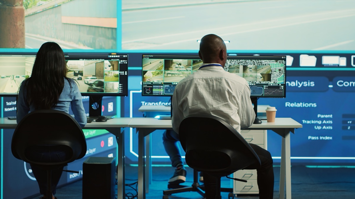 Two operators at monitoring station viewing multiple security camera feeds on large display screens