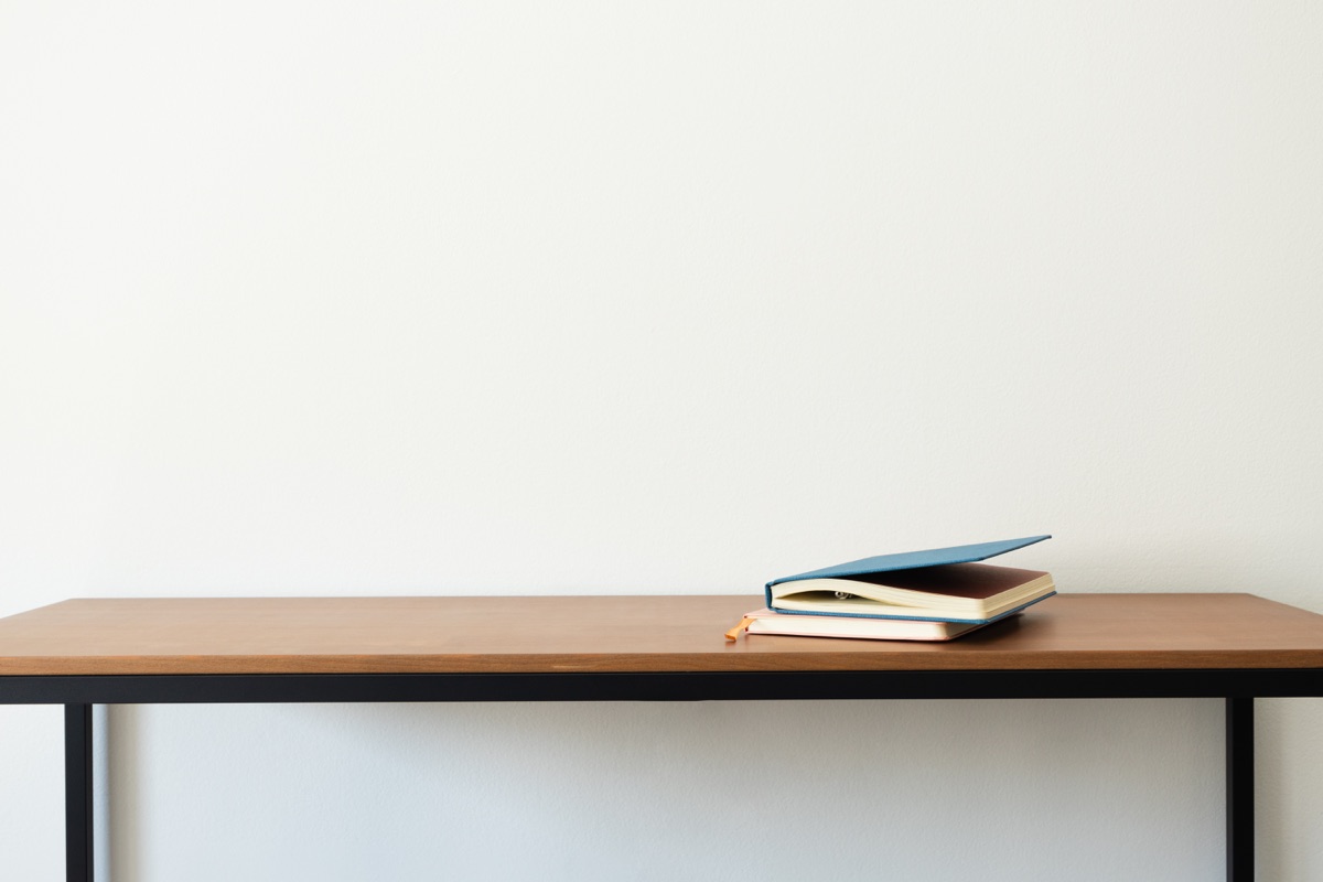 Minimalist wooden desk with black legs, two stacked notebooks against white wall